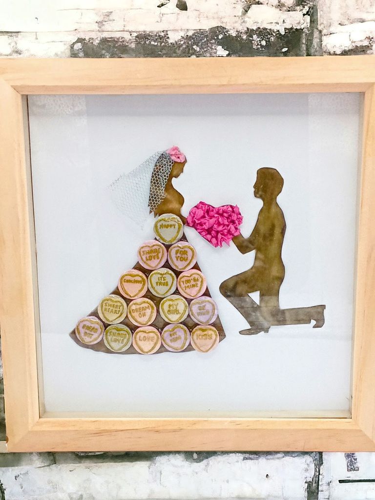 a shadow box with a resin bride and groom in the colour black gold. the brides dress is made up of sweets, in-between them is a heart made up of tissue paper 